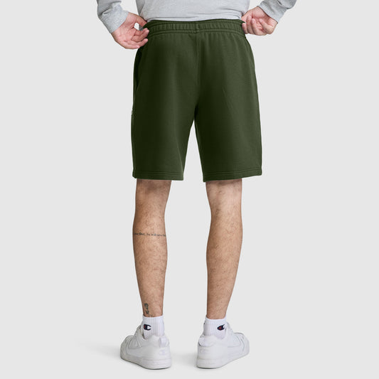 A person stands with their back to the camera, hands on hips, wearing Champion 10" Powerblend Fleece Shorts in olive green, paired with white sneakers and a light gray sweatshirt. Tattoos are visible on their lower legs.