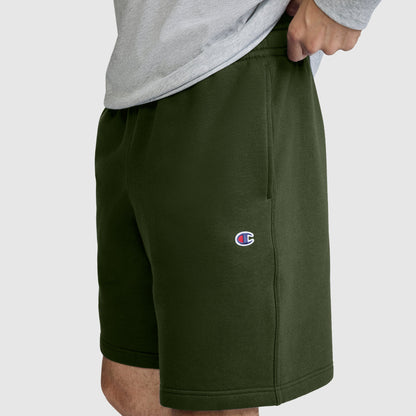 A person wearing olive green Champion 10" Powerblend Fleece Shorts and a gray shirt, with only the lower torso and upper legs visible. The Champion logo appears on the left leg of these men's shorts.