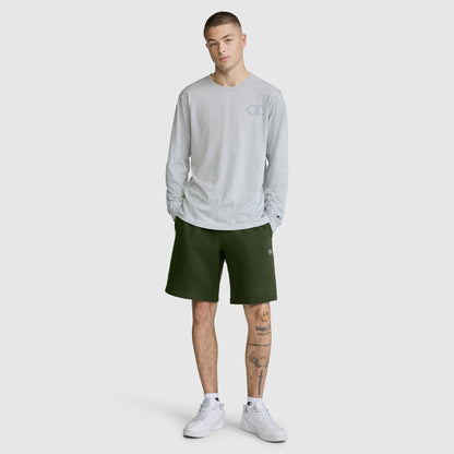 A person stands wearing a light gray long-sleeve shirt, the Champion 10" Powerblend Fleece Short in dark green, and white sneakers. They have short hair and a tattoo on their left leg. The background is plain and light-colored.