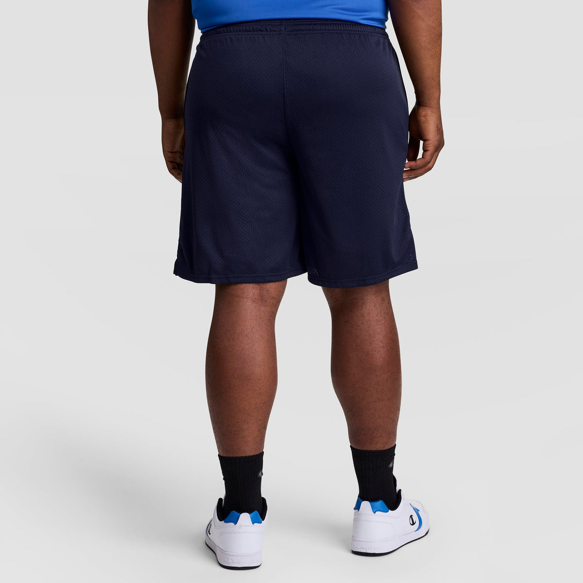 A person is shown from the back wearing Champion’s 9" Classic Mesh Shorts, C Logo - Big & Tall in navy blue, paired with a blue shirt, black socks, and white sneakers with blue accents against a plain light gray background.