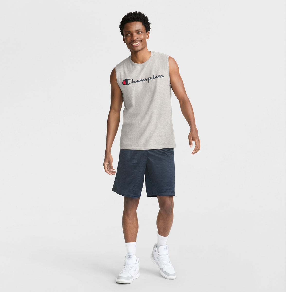 A smiling man in a sleeveless gray Champion shirt, Champion 9" Classic Mesh Shorts with C Logo in navy, white socks, and white sneakers stands against a plain white background.