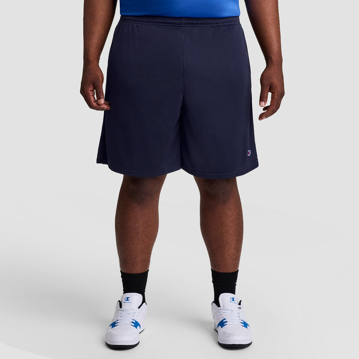 A person stands facing forward against a light gray background, showing only the lower half of their body. They are wearing Champion's 9" Classic Mesh Shorts, C Logo - Big & Tall, along with black socks and white sneakers.