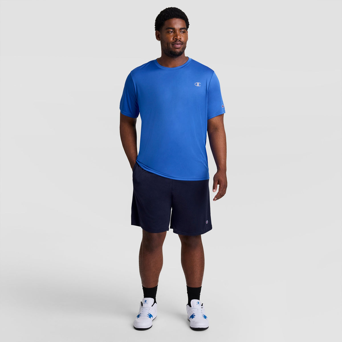 A man wearing a blue short-sleeved athletic shirt, navy 9″ Classic Mesh Shorts, C Logo - Big & Tall, and white sneakers.