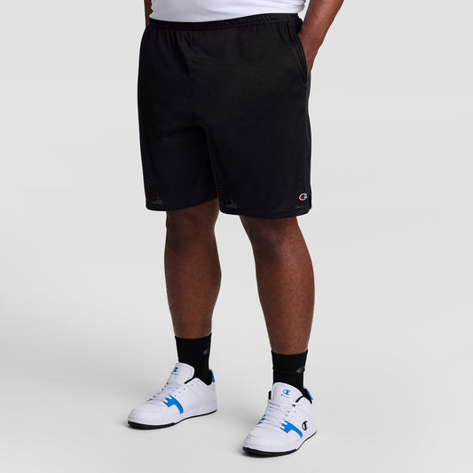 A person is shown from the waist down wearing Champion 9" Classic Mesh Shorts, C Logo - Big & Tall. They pair the black shorts with black socks and white sneakers with blue accents, standing against a plain light background.