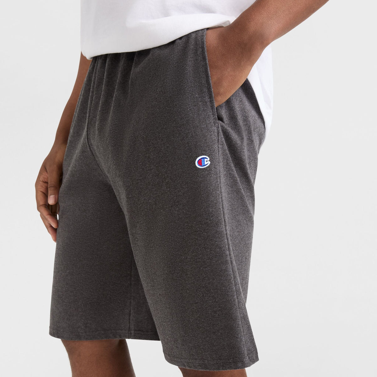 A person wearing a white shirt and Champion 9" Lightweight Lounge Shorts, C Patch Logo - Big & Tall in dark gray cotton jersey, with hands in pockets, shown from the waist down against a plain background.