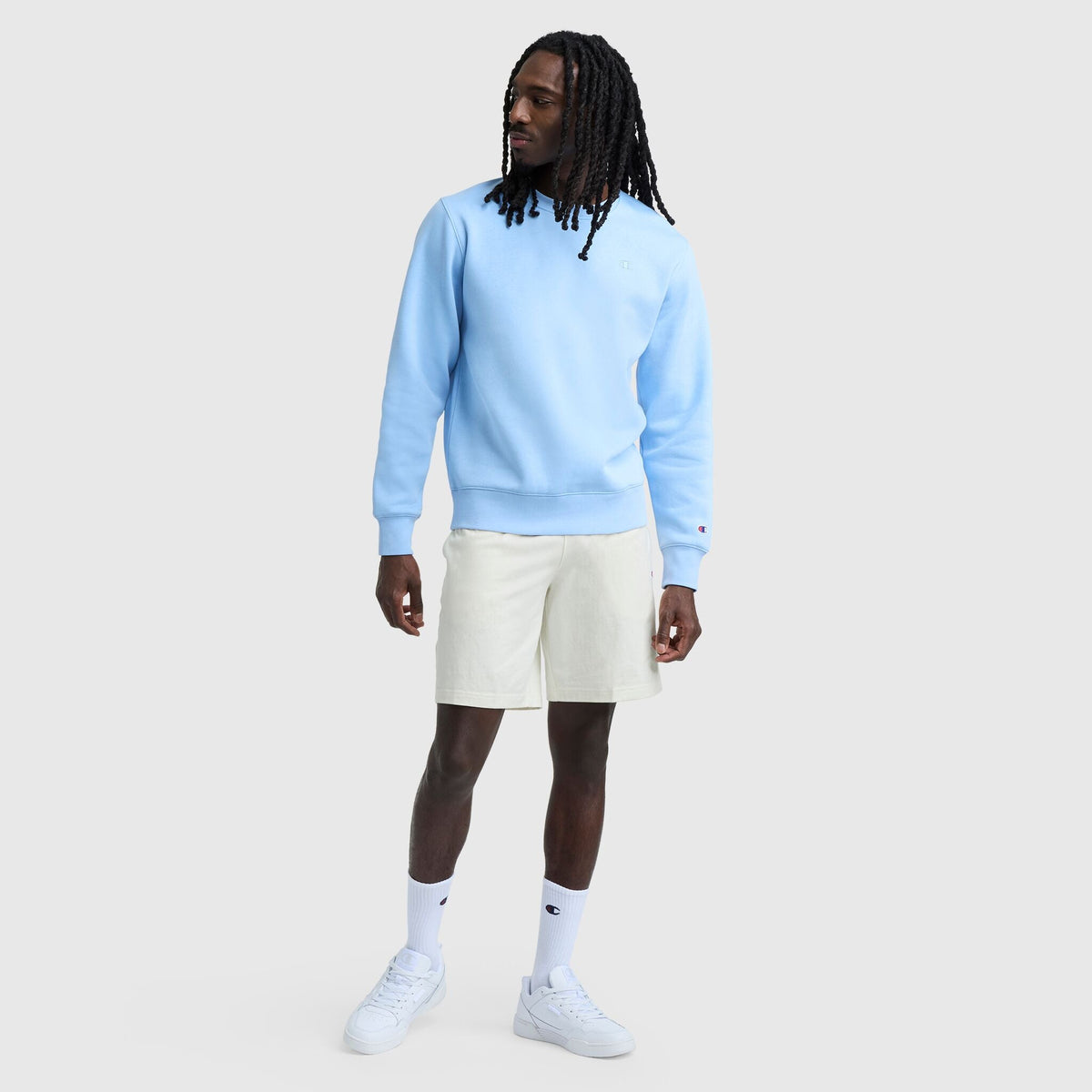 A man with long dreadlocks wears a light blue sweatshirt, Champion 9" Lightweight Lounge Shorts with C Patch Logo in off-white, white crew socks, and white sneakers, standing against a plain light gray background.