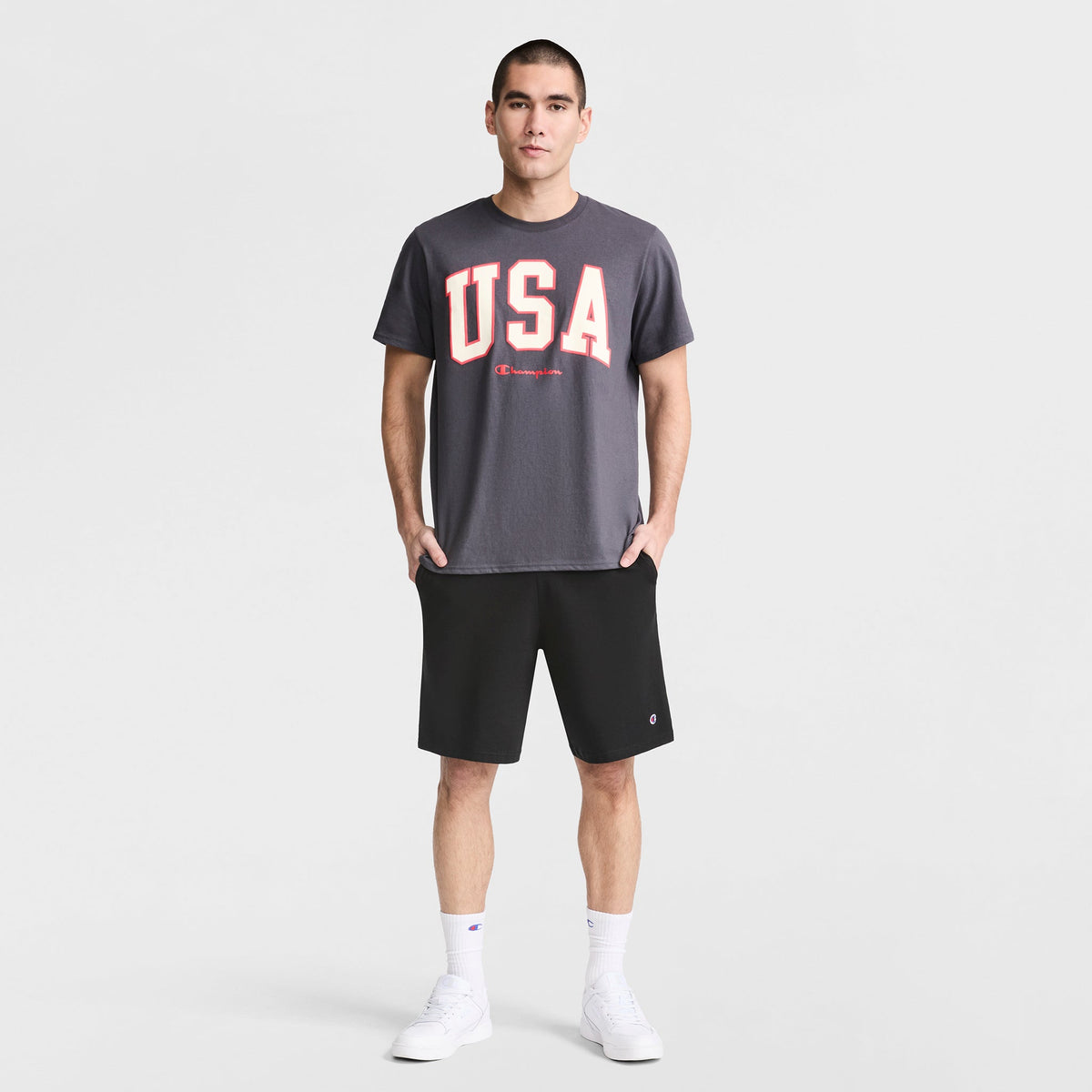 A man stands facing forward with his hands in the pockets of Champion 9" Lightweight Lounge Shorts, C Patch Logo, wearing a dark gray USA T-shirt, white crew socks, and white sneakers against a plain light background.