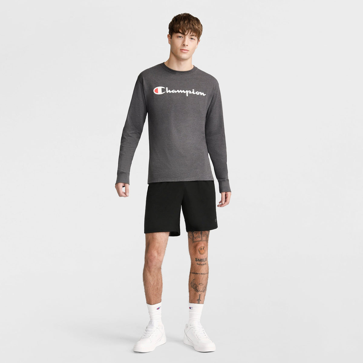 A young man stands against a plain background wearing a gray long-sleeve shirt, Champion 7" Moisture Wicking Sport Shorts in black, white crew socks, and white sneakers. He has short brown hair and tattoos on his left leg.