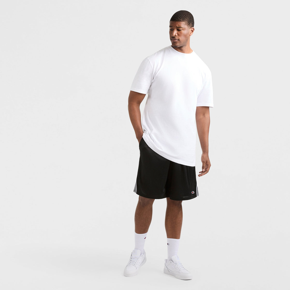 A man stands against a white background wearing a white T-shirt, Champion 10" Lacrosse Mesh Shorts - Big & Tall with vintage side taping, white crew socks, and sneakers. One hand is in his pocket as he looks down to his left.