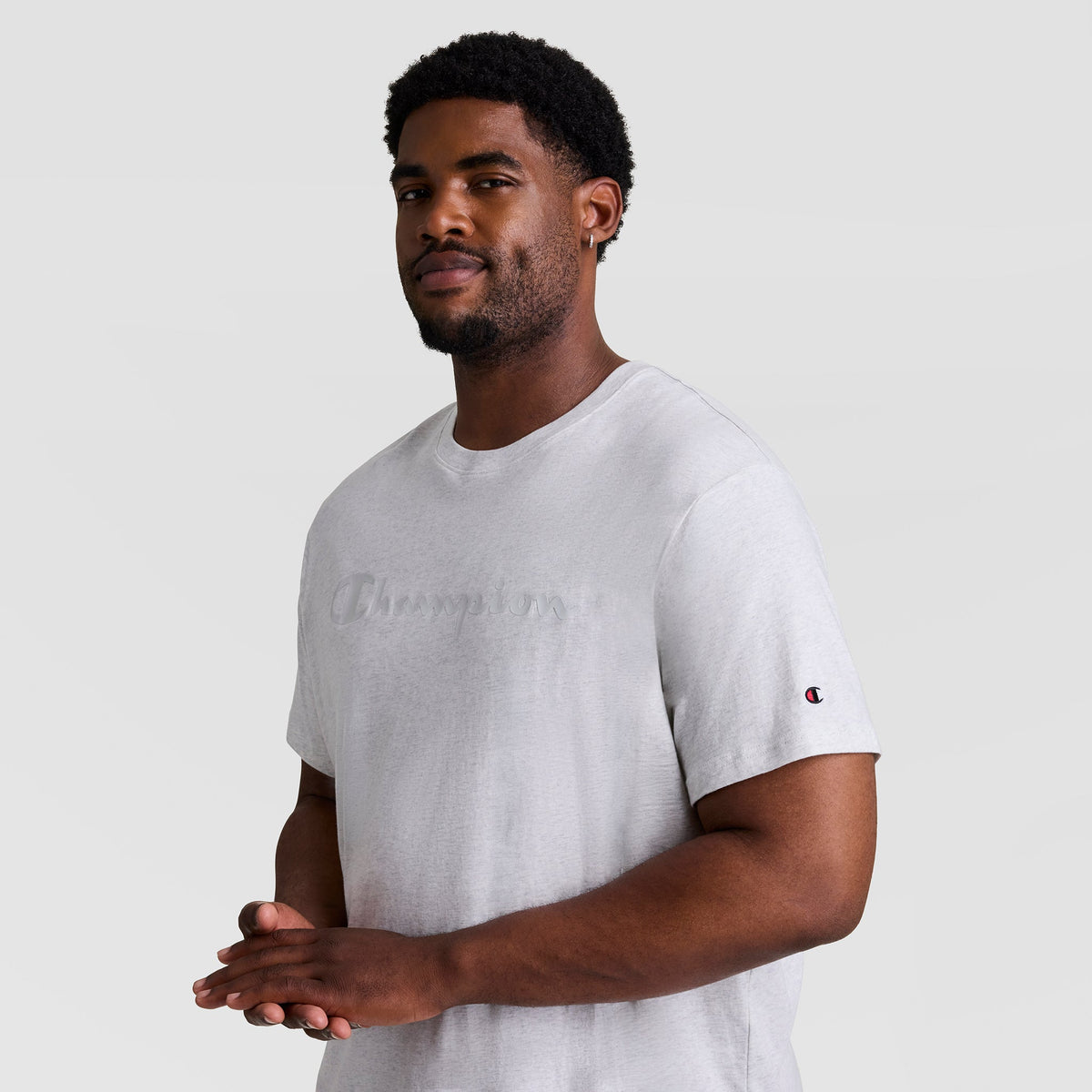 A man with short curly hair and a beard stands against a plain light background, wearing a light gray Champion Classic Graphic T-Shirt, Tonal Script Logo - Big & Tall, with the logo subtly on the front and sleeve. He looks at the camera, smiling slightly.