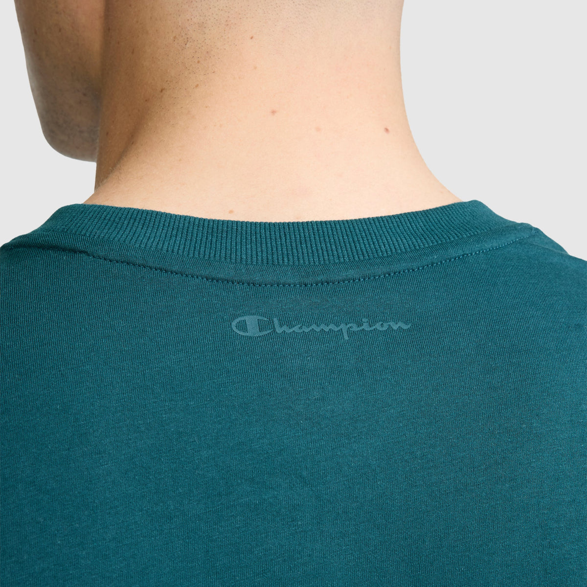 Close-up of the back of a person’s neck wearing the Champion Classic Graphic T-Shirt, Tonal Script Logo for men in teal, made from 100% cotton. The gray Champion logo is printed just below the collar, and the background is plain light gray.
