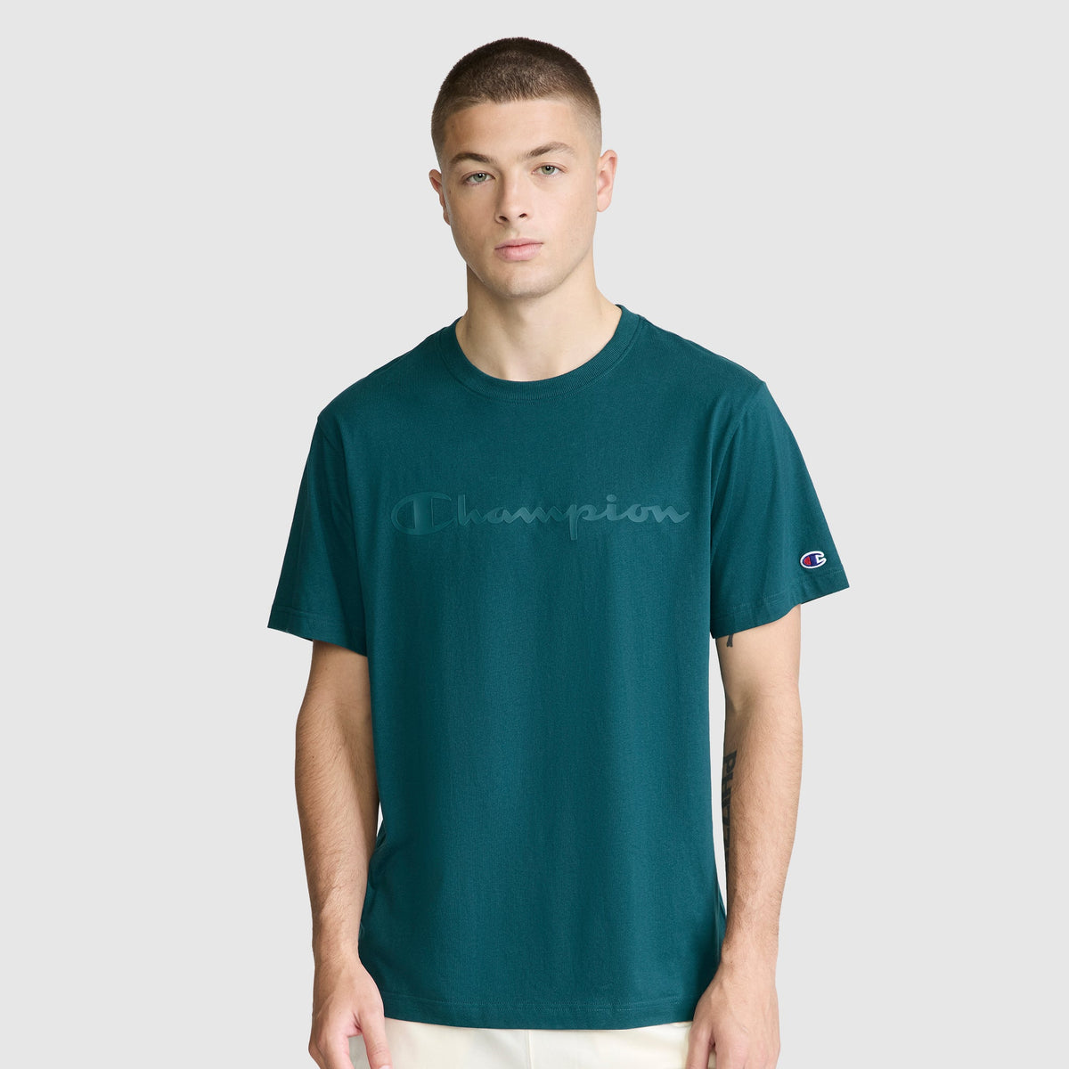 A young man wearing a teal Champion Classic Graphic T-Shirt with a tonal script logo stands against a plain light gray background, looking straight ahead with a neutral expression.