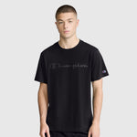 A young man wearing the Champion Classic Graphic T-Shirt with a tonal script logo stands facing forward against a plain light gray background. He has short hair and a neutral expression, highlighting the comfort of the 100% cotton fabric.