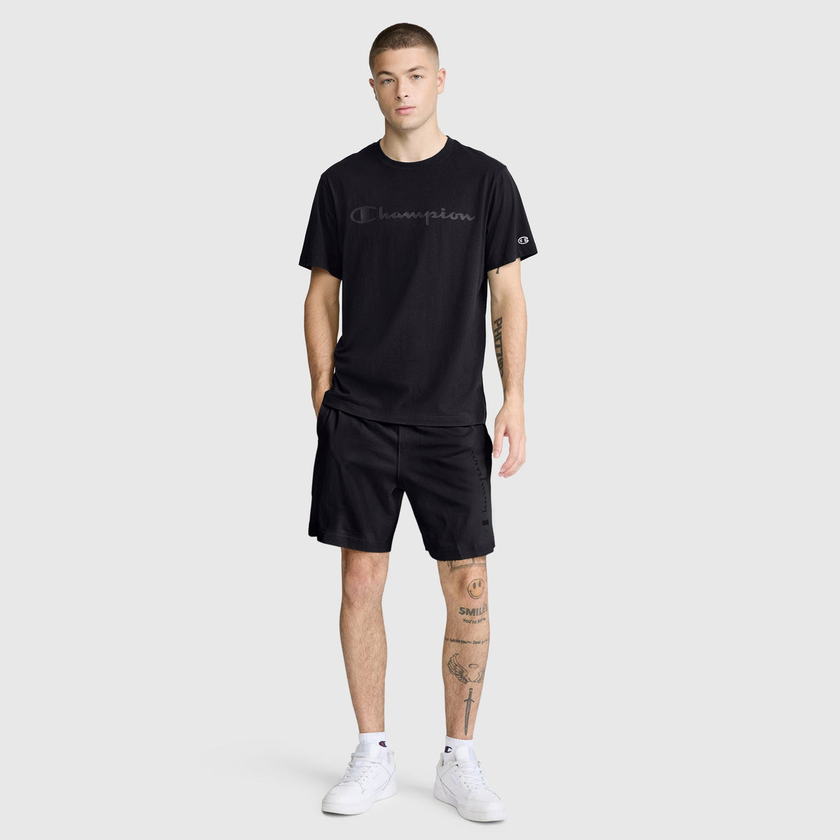 A man with short hair stands facing forward, wearing a black Champion Classic Graphic T-Shirt with a tonal script logo made from 100% cotton, paired with black shorts and white sneakers. He has tattoos on his left leg and his right hand in his pocket.