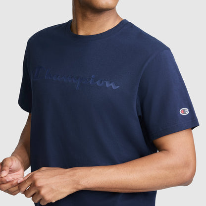 A person wearing a navy blue Champion Classic Graphic T-Shirt for men, featuring the tonal script logo on the chest and sleeve, stands against a plain light gray background.