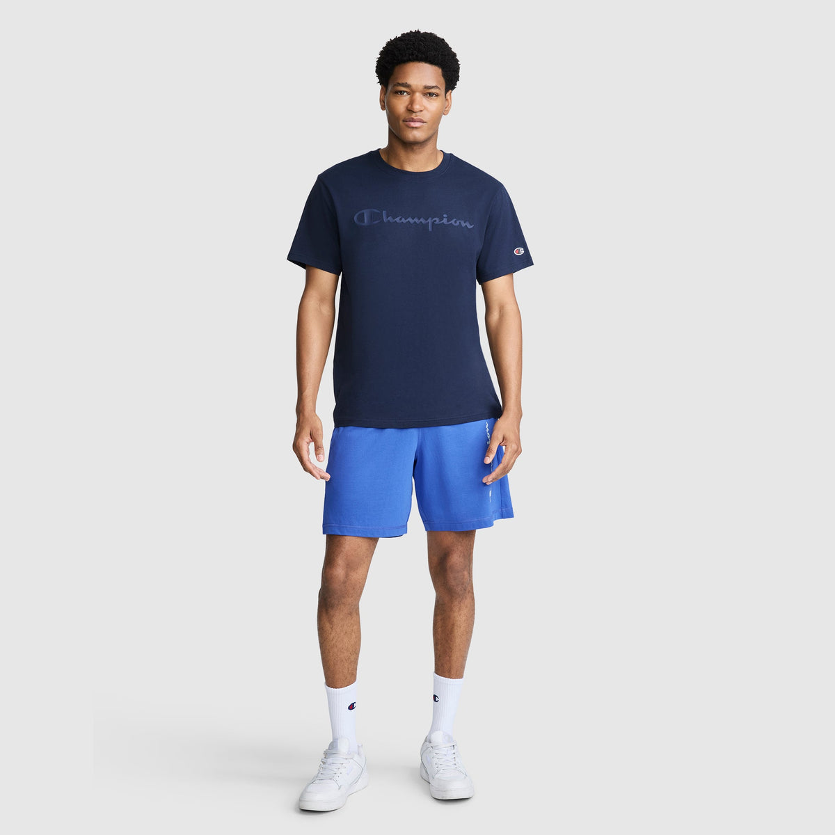A man wearing a navy Classic Graphic T-Shirt, Tonal Script Logo by Champion with a tonal logo on the chest, blue athletic shorts, white crew socks, and white sneakers.