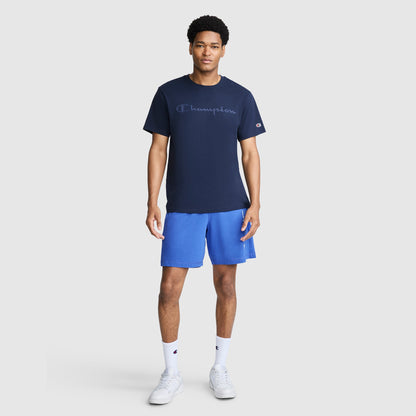 A man wearing a navy Classic Graphic T-Shirt, Tonal Script Logo by Champion with a tonal logo on the chest, blue athletic shorts, white crew socks, and white sneakers.