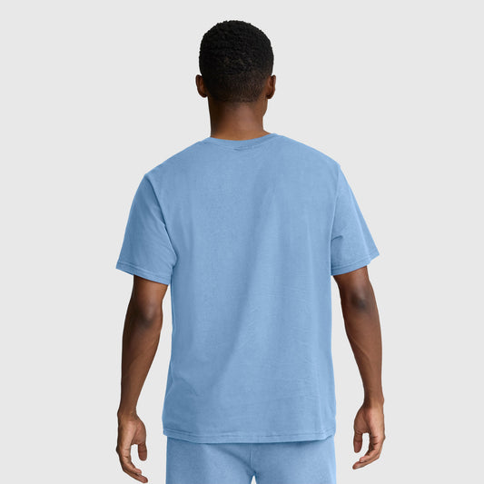 A person stands facing away from the camera, wearing a Champion Classic Graphic T-Shirt with a tonal script logo in light blue 100% cotton for men, paired with matching shorts against a plain light gray background.