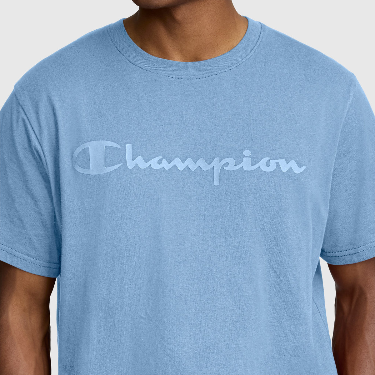 A person wearing the Champion Classic Graphic T-Shirt, Tonal Script Logo—a men’s tee made from 100% cotton in light blue, featuring “Champion” scripted across the chest in a matching blue, stylized font.