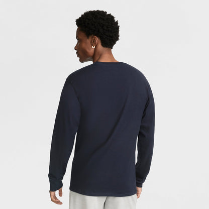 A person with short curly hair is shown from the back, wearing a Champion Classic Graphic Long-Sleeve T-Shirt, Script Logo in dark navy and light pants against a plain white background.