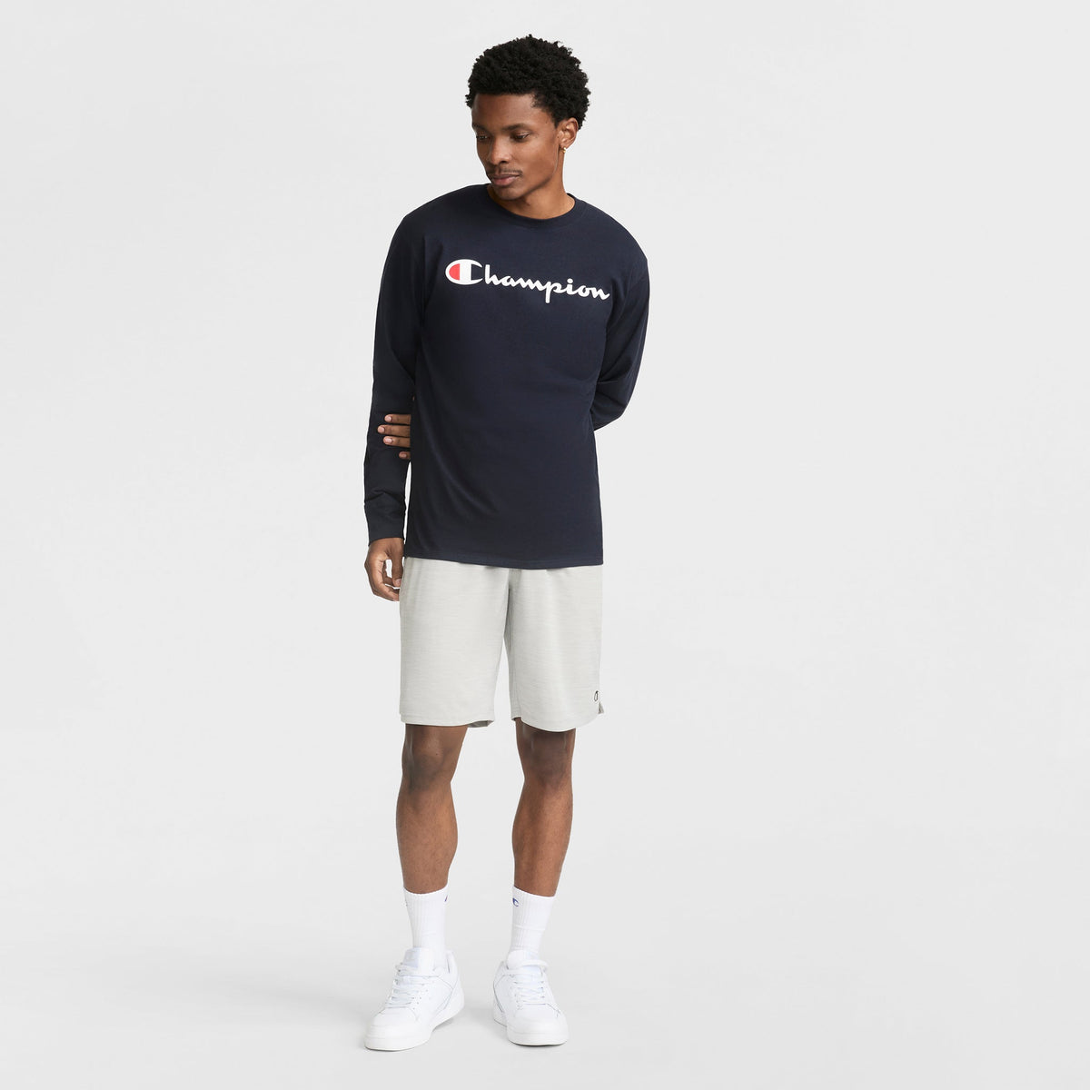 A man stands against a white background, wearing the Champion Classic Graphic Long-Sleeve T-Shirt with Script Logo in navy, paired with light gray shorts, white socks, and white sneakers. He looks downward with one arm behind his back.