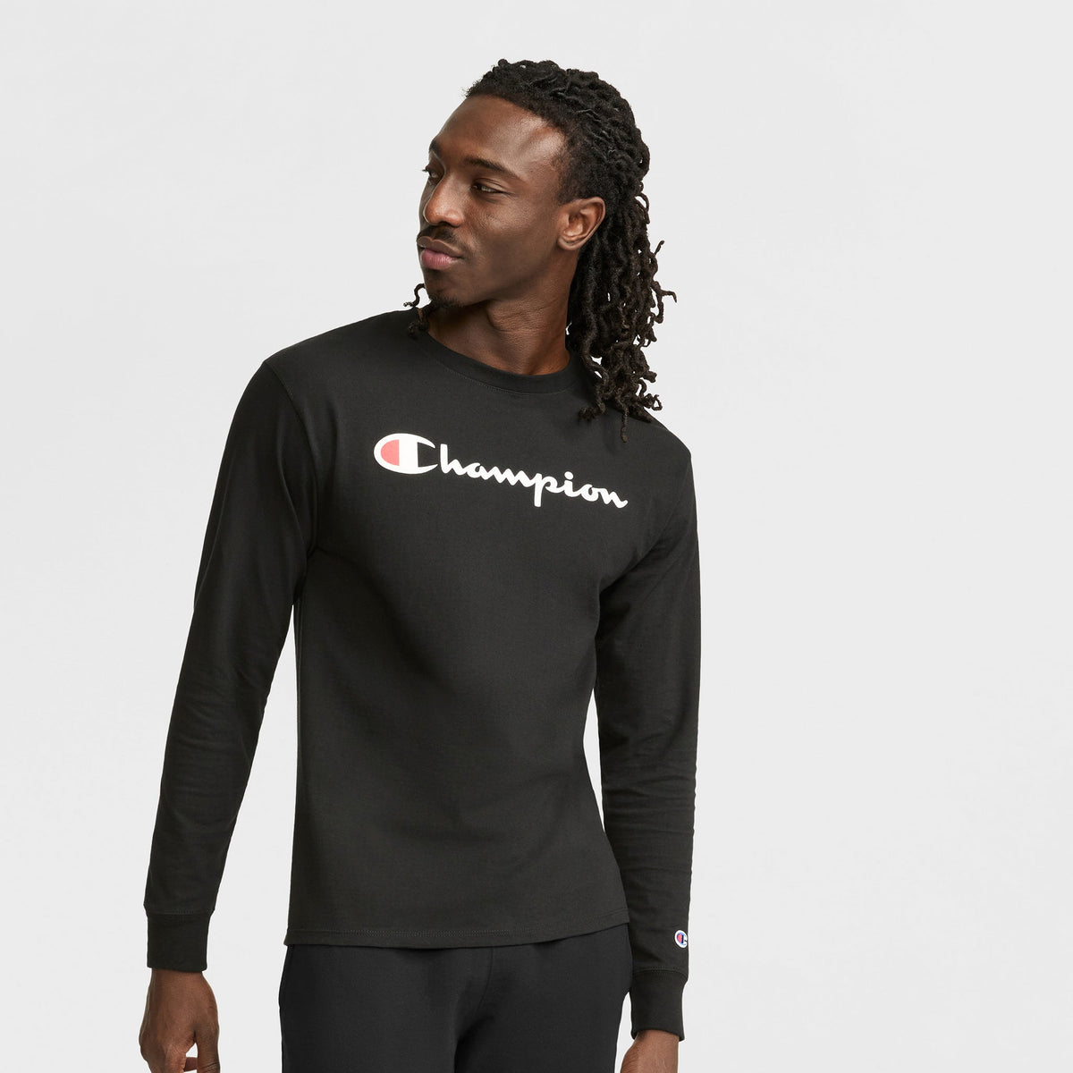 A man with long dreadlocks wears a black Champion Classic Graphic Long-Sleeve T-Shirt with a script logo and black pants, looking to the side with a neutral expression against a plain white background.