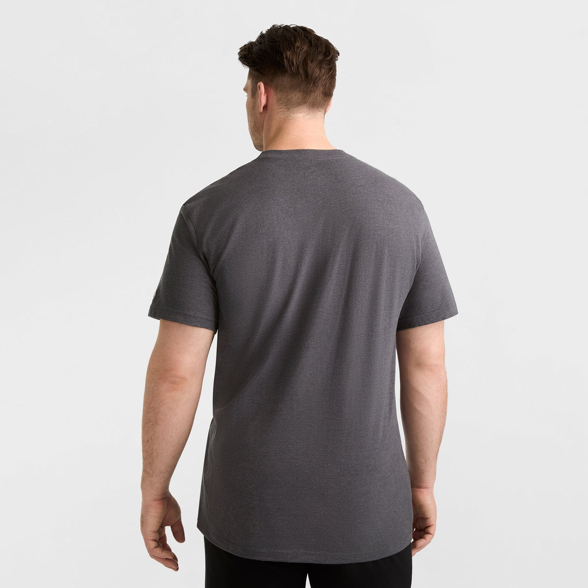 A man with short brown hair, back to the camera, wears a Champion Classic Graphic T-Shirt with Script Logo (Big & Tall) in dark gray and black pants, styled for casual streetwear against a plain light gray background.