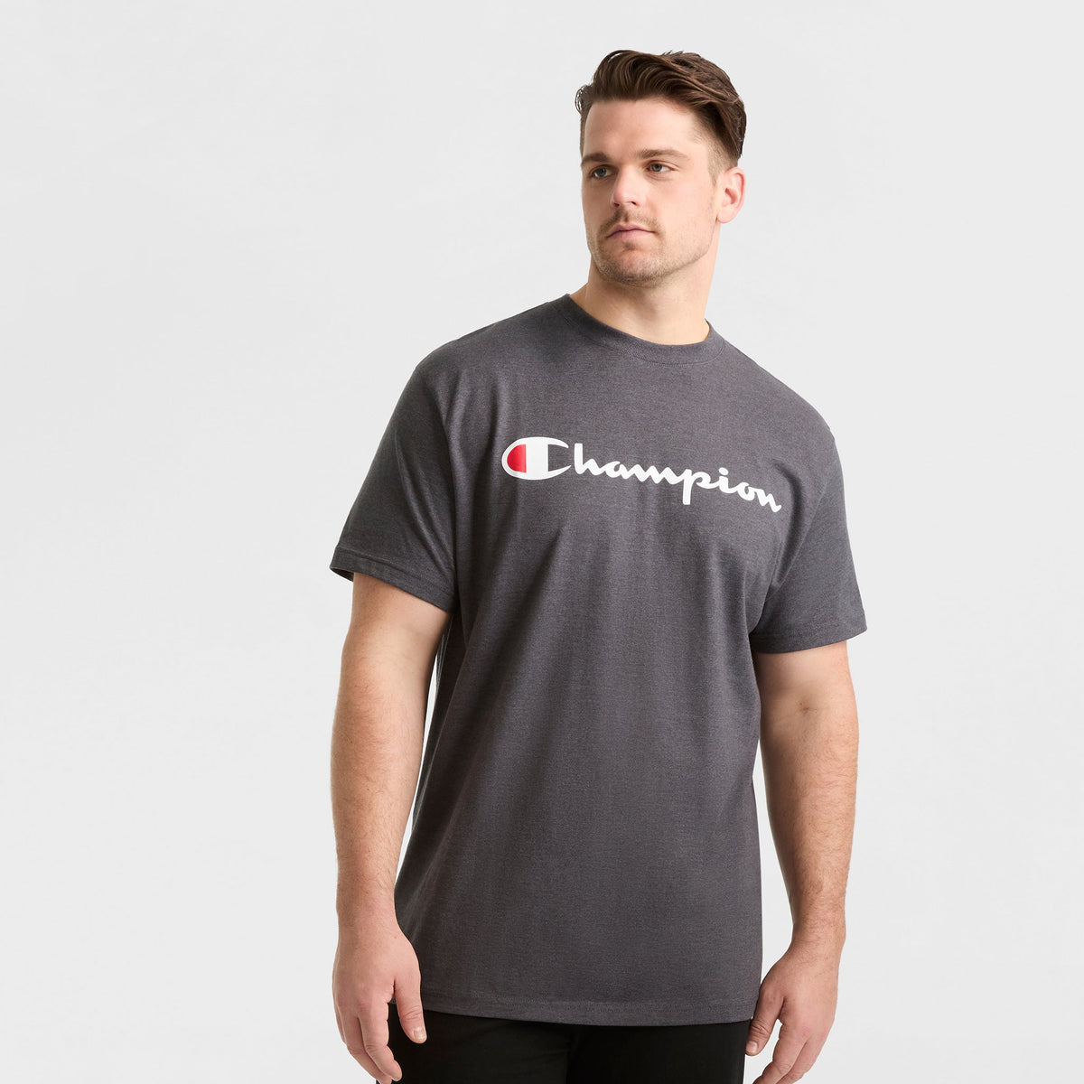 A man wears a Champion Classic Graphic T-Shirt, Script Logo - Big & Tall in dark gray, featuring the white and red Champion logo across the chest, standing against a plain light gray background.