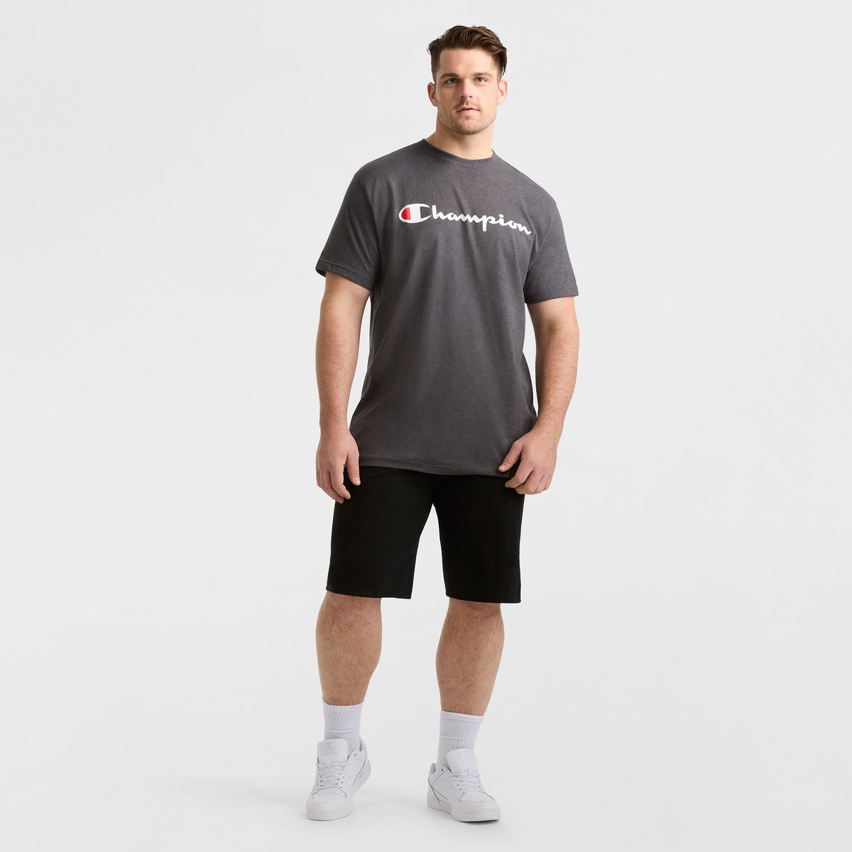 A man models a dark gray Champion Classic Graphic T-Shirt, Script Logo - Big & Tall, paired with black shorts, white socks, and white sneakers against a plain white background for a classic streetwear look.