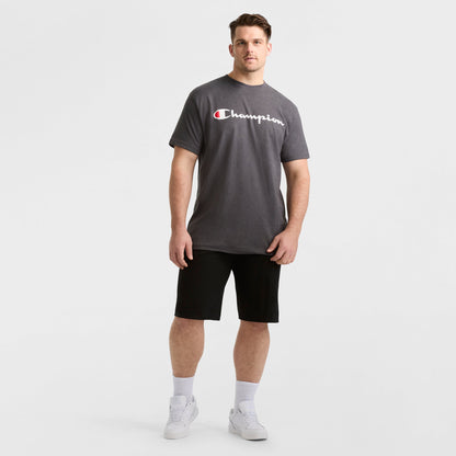 A man models a dark gray Champion Classic Graphic T-Shirt, Script Logo - Big & Tall, paired with black shorts, white socks, and white sneakers against a plain white background for a classic streetwear look.