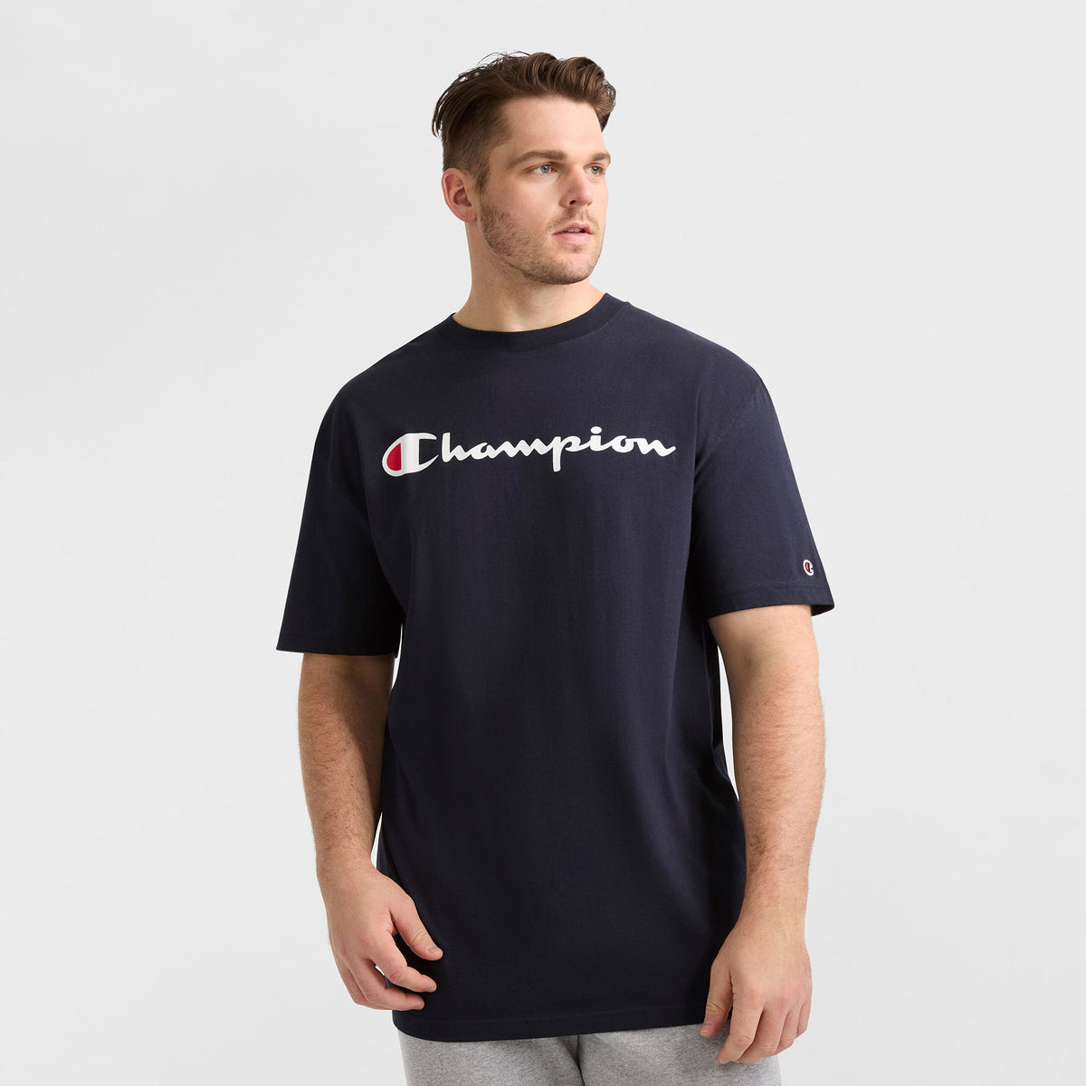 A man in a loose-fitting Champion Classic Graphic T-Shirt, Script Logo - Big & Tall, with white logo text and light gray pants stands against a white background, showcasing effortless streetwear style as he looks left.