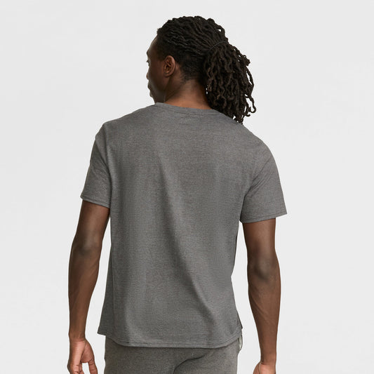 A person with long, dark, braided hair stands with their back to the camera, wearing a Champion Classic Graphic T-Shirt with a script logo and gray pants in a casual streetwear style against a white background.
