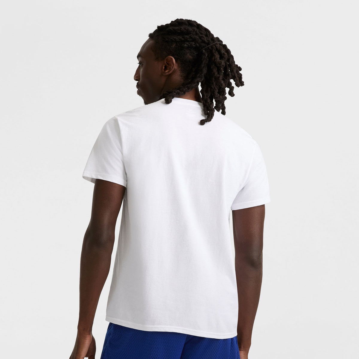 A person with dark, braided hair faces away from the camera, wearing a Champion Classic Graphic T-Shirt with Script Logo and blue shorts, creating a simple yet classic streetwear look against a plain white background.