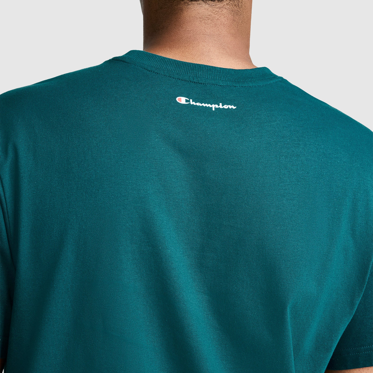 A person is shown from the back wearing a teal Champion Classic Graphic T-Shirt, Script Logo, made from 100% cotton. The small Champion logo appears near the neckline of the shirt.