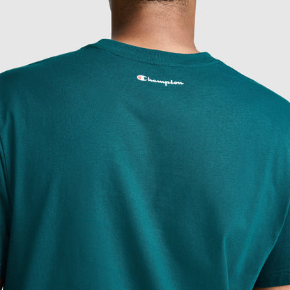 A person is shown from the back wearing a teal Champion Classic Graphic T-Shirt, Script Logo, made from 100% cotton. The small Champion logo appears near the neckline of the shirt.
