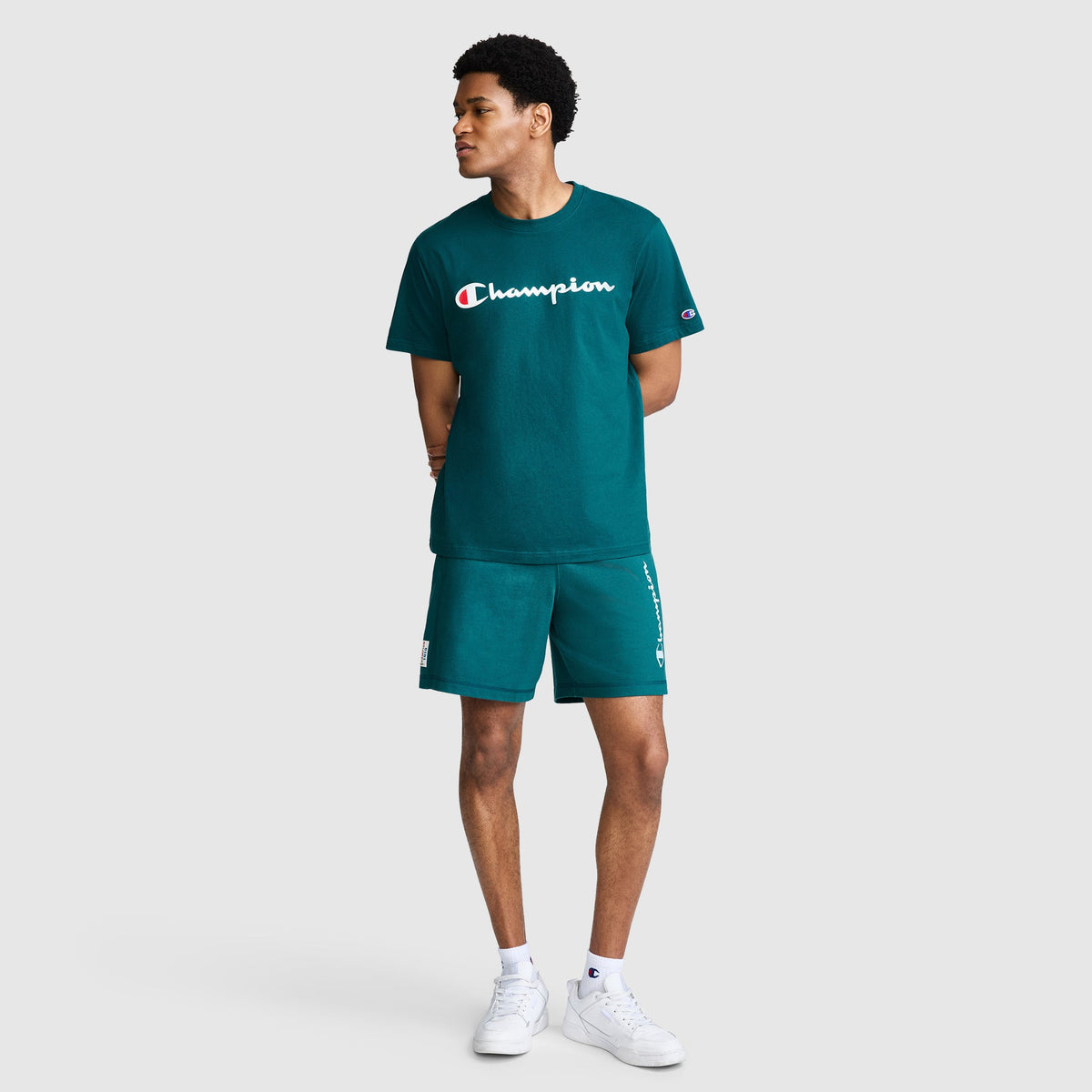 A person wearing the Champion Classic Graphic T-Shirt, Script Logo in teal, paired with matching shorts, stands with hands behind their back and looks to the side. They are also wearing white sneakers against a plain light gray background.