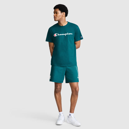 A person wearing the Champion Classic Graphic T-Shirt, Script Logo in teal, paired with matching shorts, stands with hands behind their back and looks to the side. They are also wearing white sneakers against a plain light gray background.