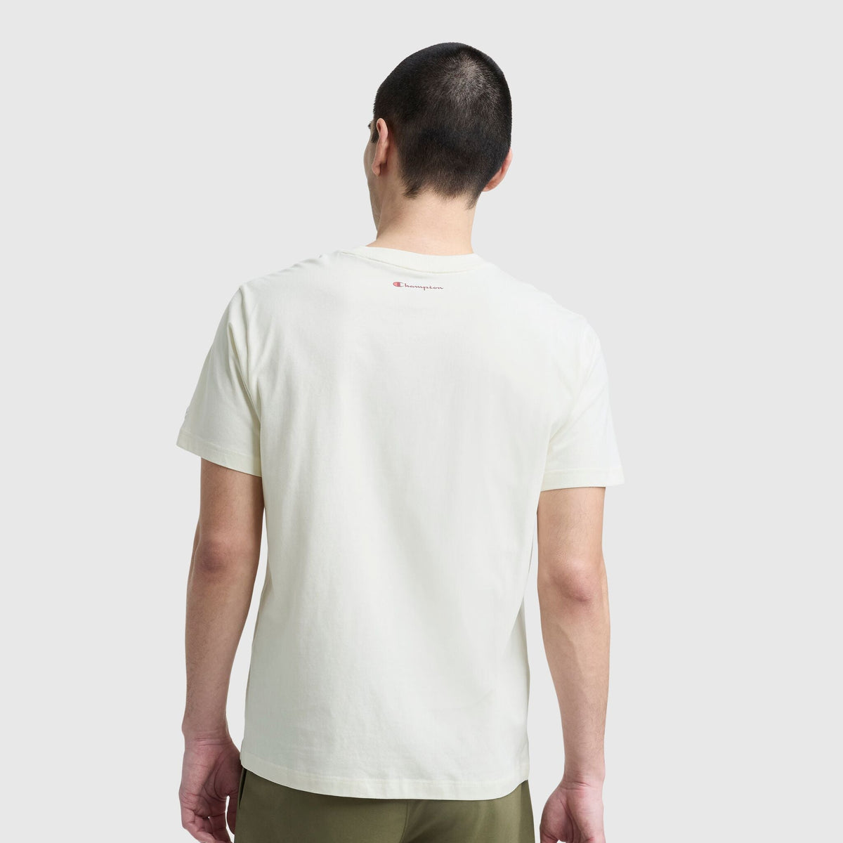 A person with short dark hair is seen from behind wearing a plain white Champion Classic Graphic T-Shirt, Script Logo, and olive green pants against a light gray background.