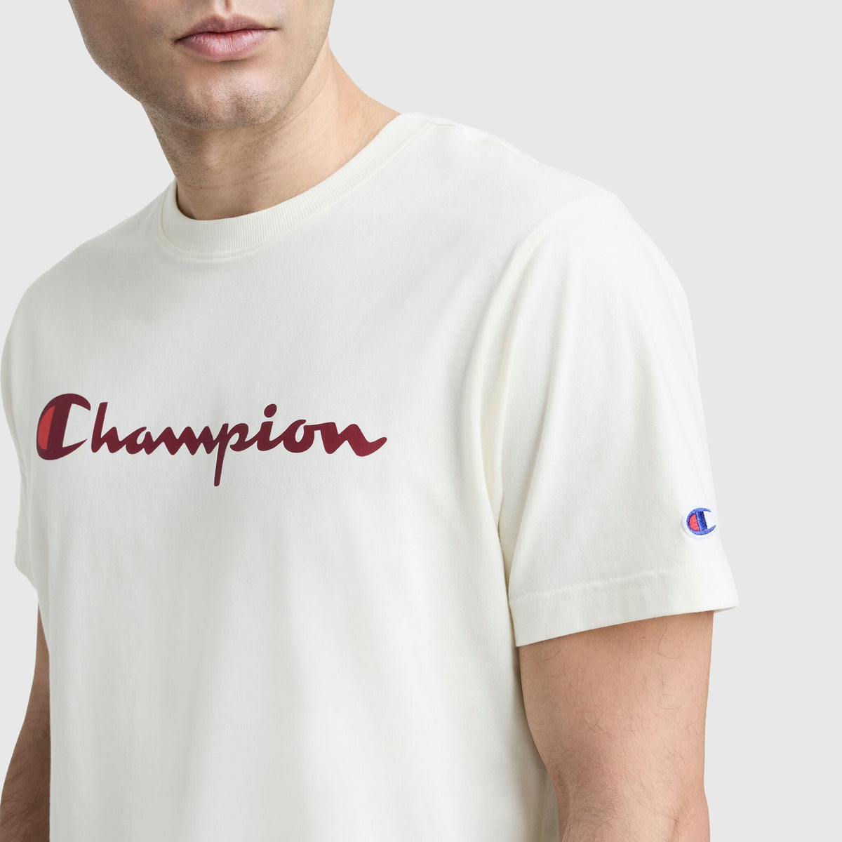 A person wearing a Champion Classic Graphic T-Shirt, Script Logo—an off-white streetwear staple featuring the red Champion script across the chest and a small sleeve emblem—shown from shoulders to mid-arm.