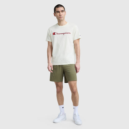 A man faces forward in a white Champion Classic Graphic T-Shirt with Script Logo, olive green shorts, white crew socks, and white sneakers—creating a relaxed streetwear vibe against a simple light gray background.