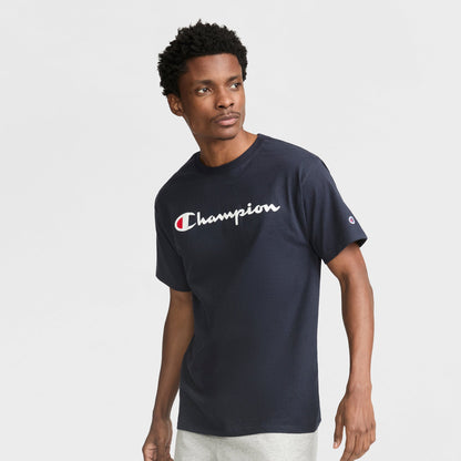 A man wears the Champion Classic Graphic T-Shirt with white script logo, paired with light gray shorts, as he stands against a plain white background and looks slightly to the side.
