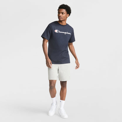 A man stands against a white backdrop in a navy Champion Classic Graphic T-Shirt with Script Logo, paired with light gray shorts, white crew socks, and sneakers—capturing an easy streetwear look as he glances sideways with one arm slightly raised.