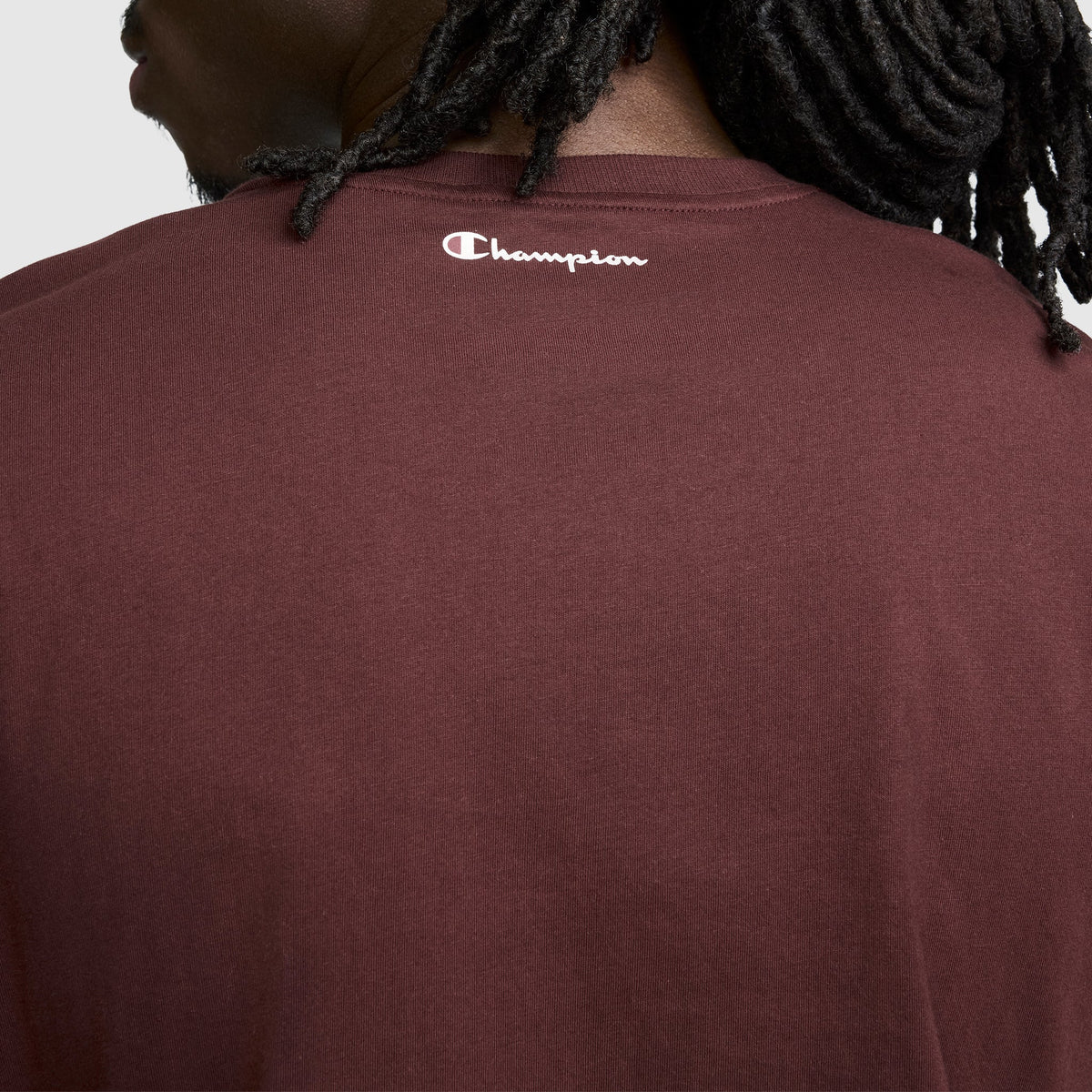 A person with dark, twisted hair wears the Champion Classic Graphic T-Shirt, Script Logo—a maroon men’s tee that perfectly embodies streetwear style. The back of this shirt displays a small white script logo near the neckline.