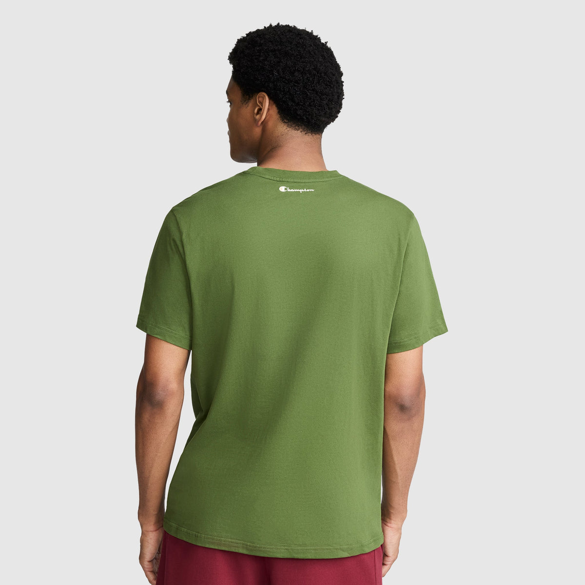 A person wearing a green Champion Classic Graphic T-Shirt with the Script Logo and red shorts stands facing away, showcasing the back of the shirt against a plain gray background—a simple yet classic streetwear look.