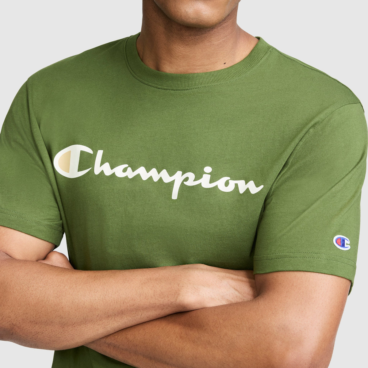 A person wearing the Champion Classic Graphic T-Shirt, Script Logo in green, featuring the signature Champion script across the chest and a small emblem on the sleeve. Channeling classic streetwear style, their arms are crossed and their face is partially obscured.