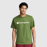 A man wearing the Champion Classic Graphic T-Shirt, Script Logo in green—with "Champion" displayed across the chest in bold white letters—stands against a plain light gray background, embodying timeless streetwear style.
