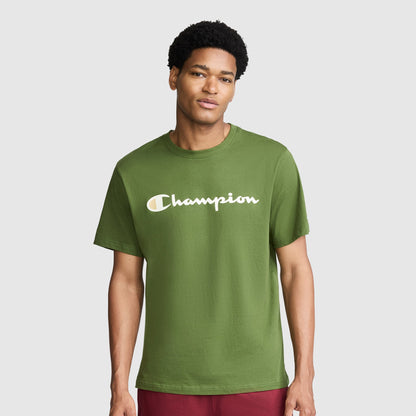 A man wearing the Champion Classic Graphic T-Shirt, Script Logo in green—with "Champion" displayed across the chest in bold white letters—stands against a plain light gray background, embodying timeless streetwear style.