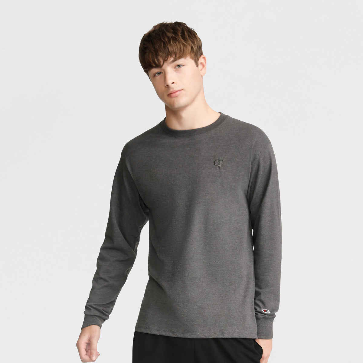 A young man with short brown hair wears a Champion Classic Long Sleeve T-Shirt with embroidered C logo in dark gray and black pants, standing against a light gray background.