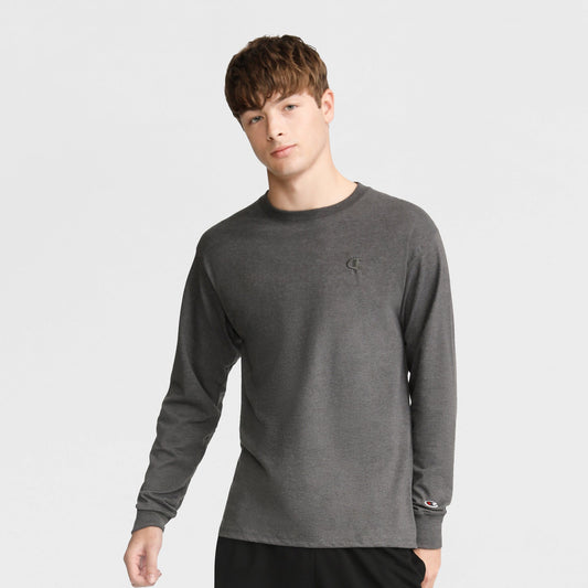 A young man with short brown hair wears a Champion Classic Long Sleeve T-Shirt with embroidered C logo in dark gray and black pants, standing against a light gray background.