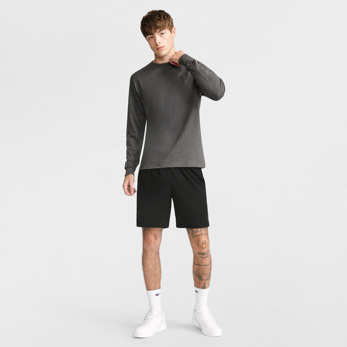 A young man with tattoos on his left leg wears a Champion Classic Long Sleeve T-Shirt with an embroidered C logo, paired with black shorts and white high-top sneakers, standing against a plain light background and touching his neck.